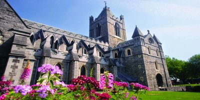 Tourism Ireland, Christ Church Angled view of brick grey castle surrounded by vibrant coloured pink and purple flowers sitting on a bed of bright green grass