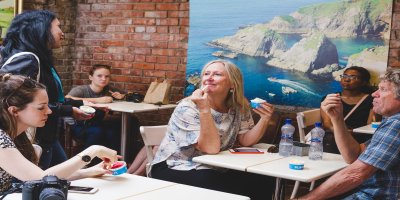 inside by delicious dublin Red brick wall in the background with a large poster of the blue sea and cliffs handing on the wall. Four white tables can be seen with people sitting at them tasting food.