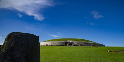 Tourism Ireland, Newgrange Bright blue skies with small white fluffy cloud in the top left corner. Bright Green grass with stone and grass historic site on the hill.