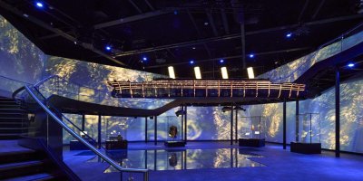 titanic-belfast-experience-tourism-ireland-photgraphed-by-richard-watson Blue light illusions projected on the 3 back walls. stairs with silver hand rail from the left foreground all the way around the room. 5 clear glass boxes with artefacts on the ground in the middle of the room.
